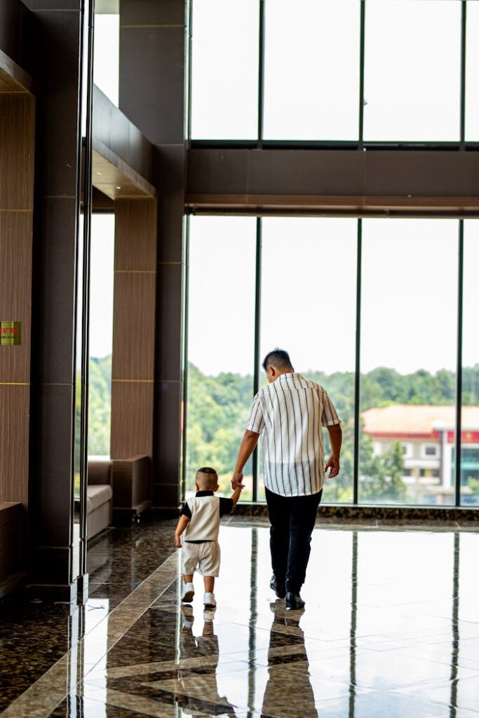 A father and child walk hand in hand in a modern building with large windows.